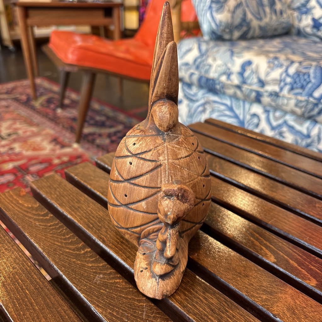 A wooden pineapple and small sitting figure toothpick holder on a wooden bench in a room setting.