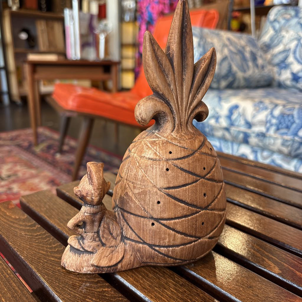 A wooden pineapple and small sitting figure toothpick holder on a wooden bench in a room setting.