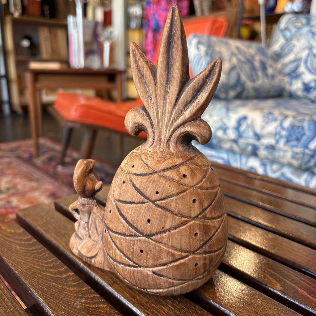 A wooden pineapple and small sitting figure toothpick holder on a wooden bench in a room setting.