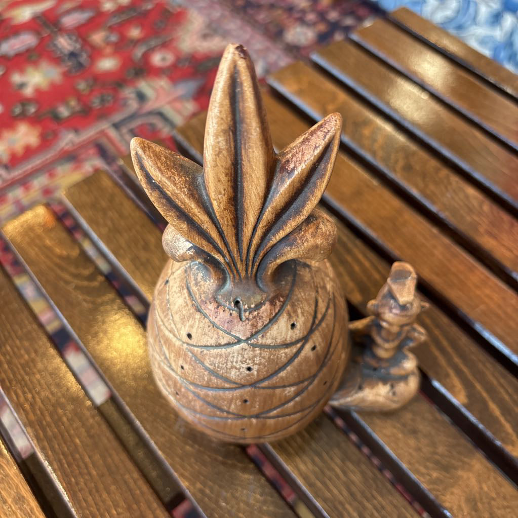A wooden pineapple and small sitting figure toothpick holder on a wooden bench in a room setting.