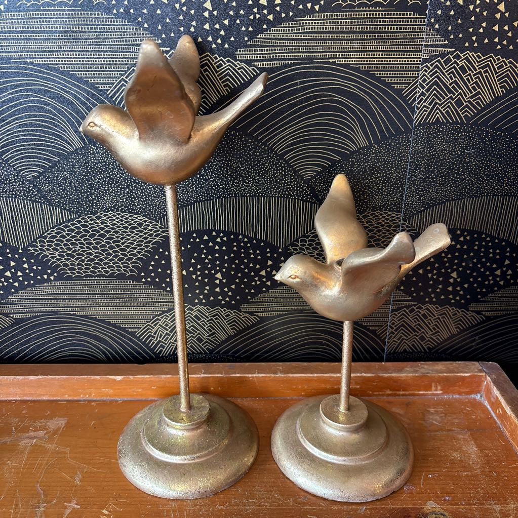 Two vintage-style brass bird figurines mounted on slender stands with round bases, displayed on a wooden surface against a dark patterned wallpaper background.