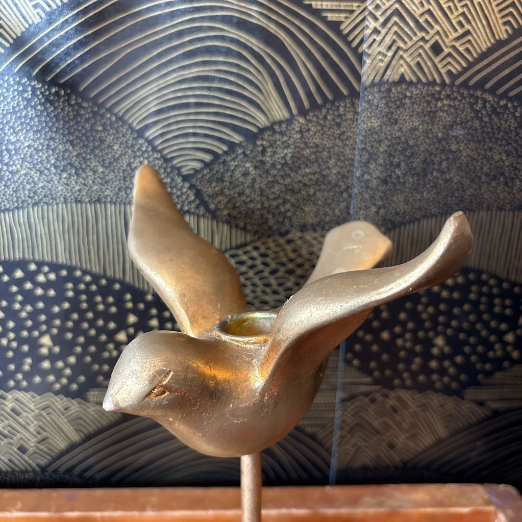 A small metallic bird figurine with wings spread, mounted on a thin stand, photographed against a patterned dark blue and beige background.