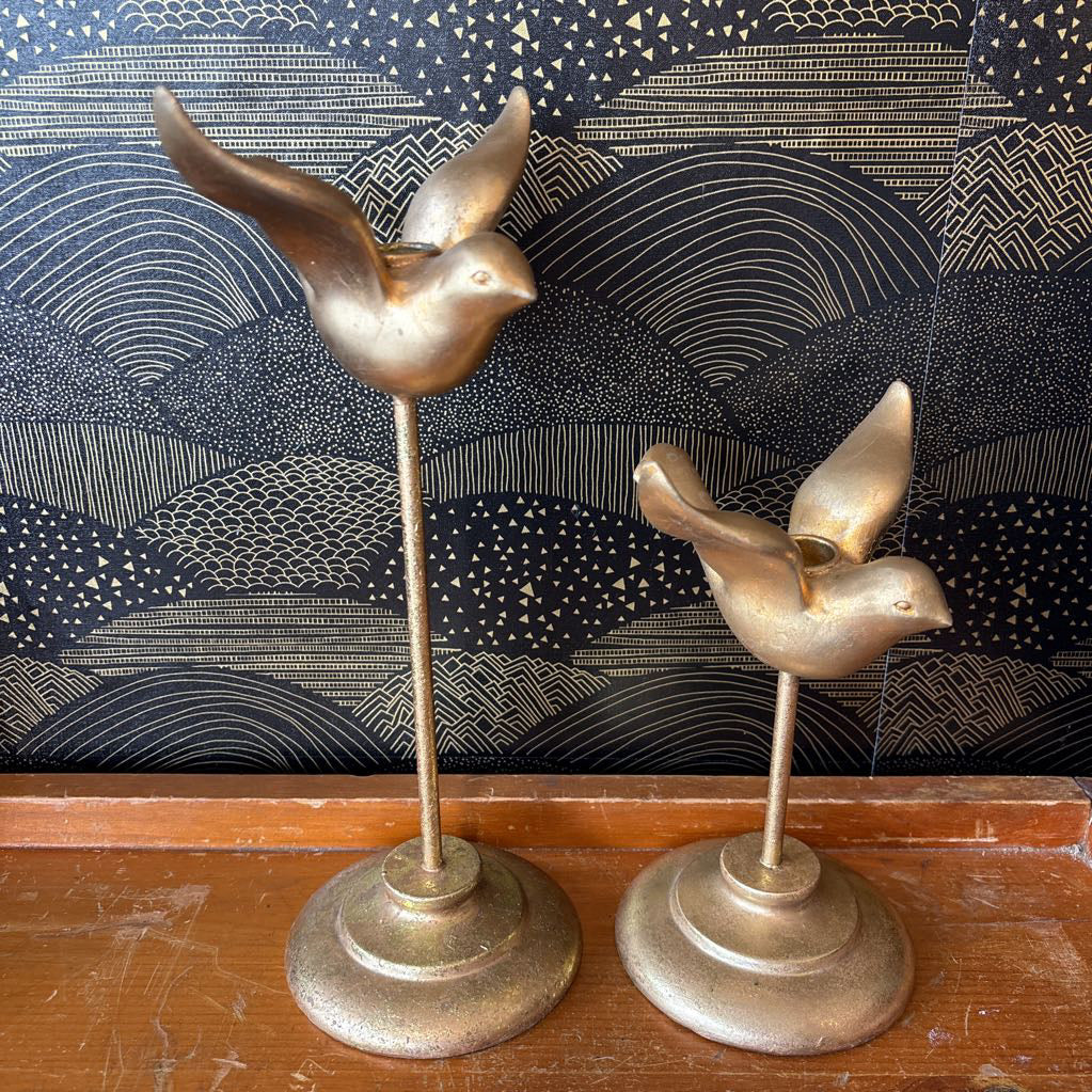 Two vintage brass bird candlesticks on a wooden surface against a patterned black and gold background.