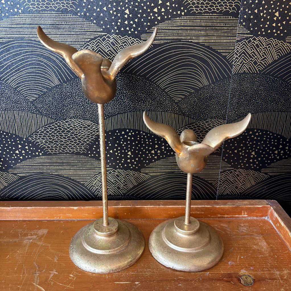 Two vintage brass bird-shaped candle holders with outstretched wings on slender stems and round bases, displayed on a wooden surface against a dark patterned backdrop.
