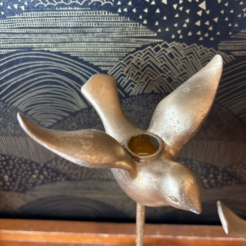 A vintage-style metal bird-shaped candle holder with wings spread, mounted on a thin metal stand. The bird has a weathered metallic finish with a small glass cup inside its neck.