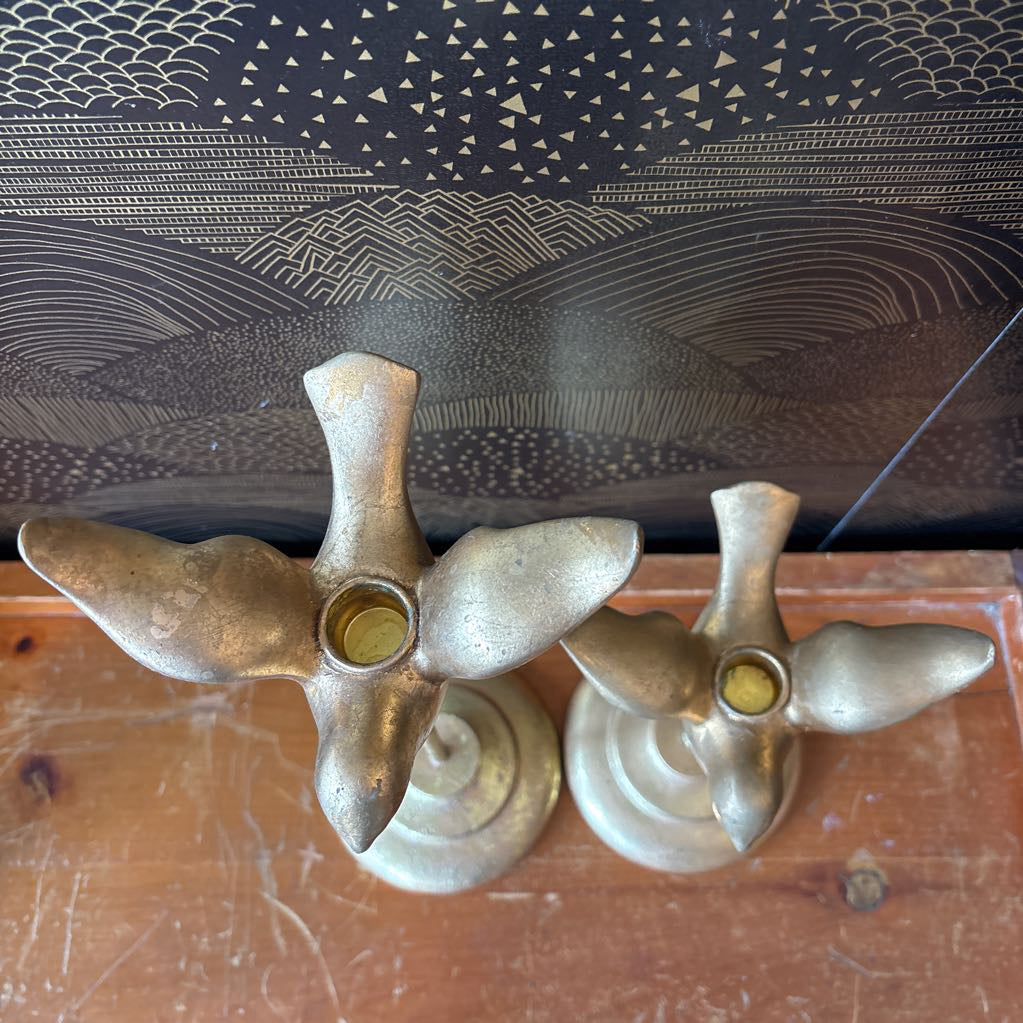 A pair of vintage brass bird-shaped candle holders with a polished metallic finish, resting on a wooden surface in front of a patterned black and gold backdrop.