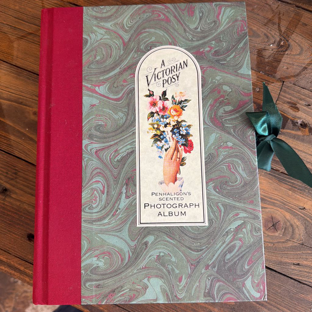 A vintage-style photo album titled "A Victorian Posy" lies on a wooden surface. The album has a red spine and a marbled cover in muted green, gray, and pink tones, with a central label showing a hand holding flowers.