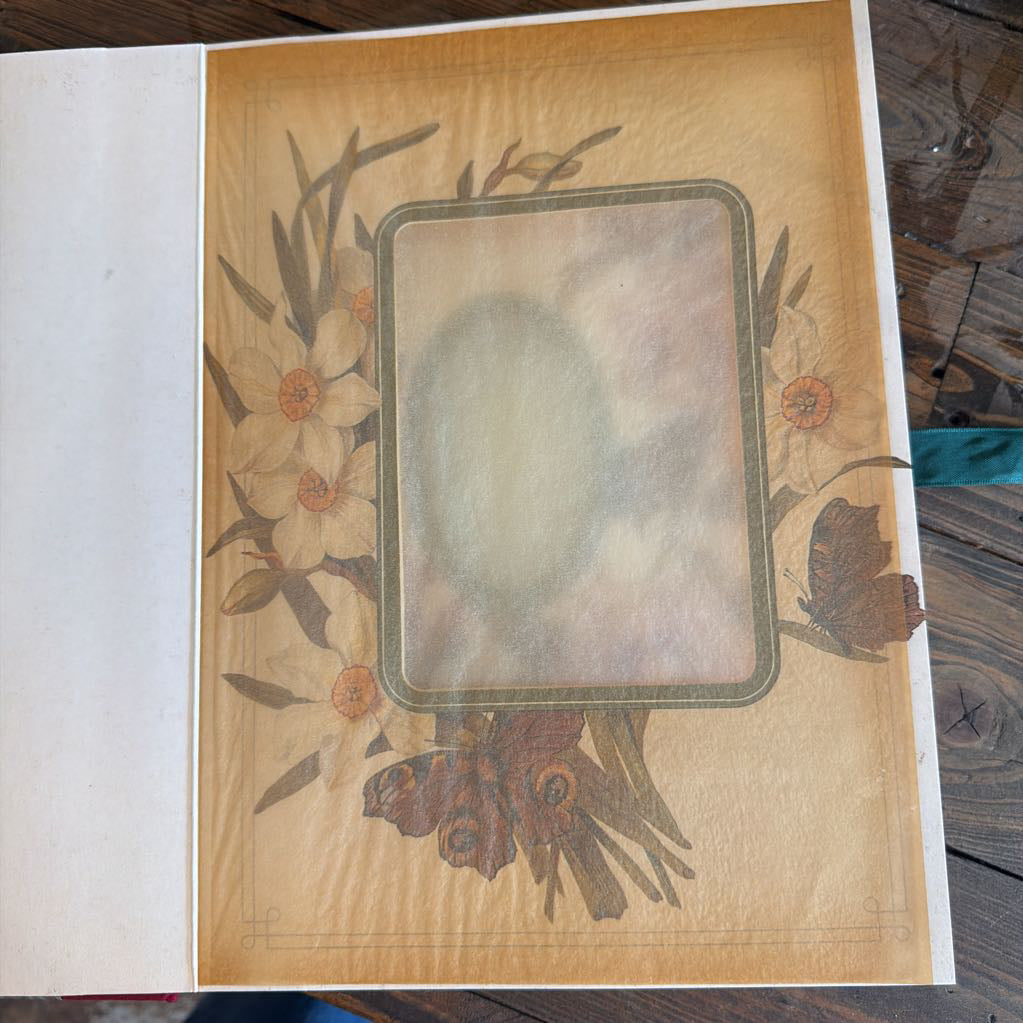 A vintage-style printed card or page with a tan background, floral border illustration, and a central rounded rectangular photo slot or blank area, photographed on a wooden surface.