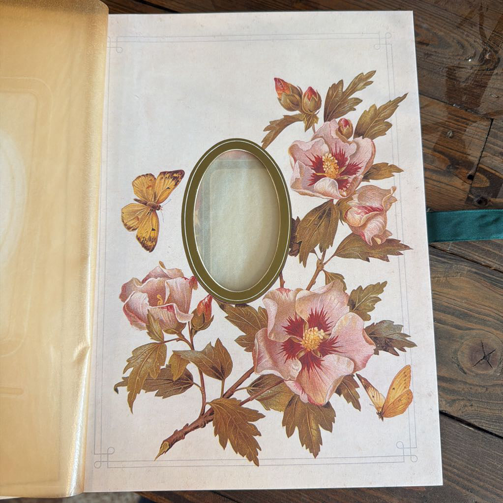 An antique oval mirror with a gold-toned frame, mounted on a vintage floral page insert featuring pink hibiscus flowers and butterflies, displayed on a rustic wooden table.