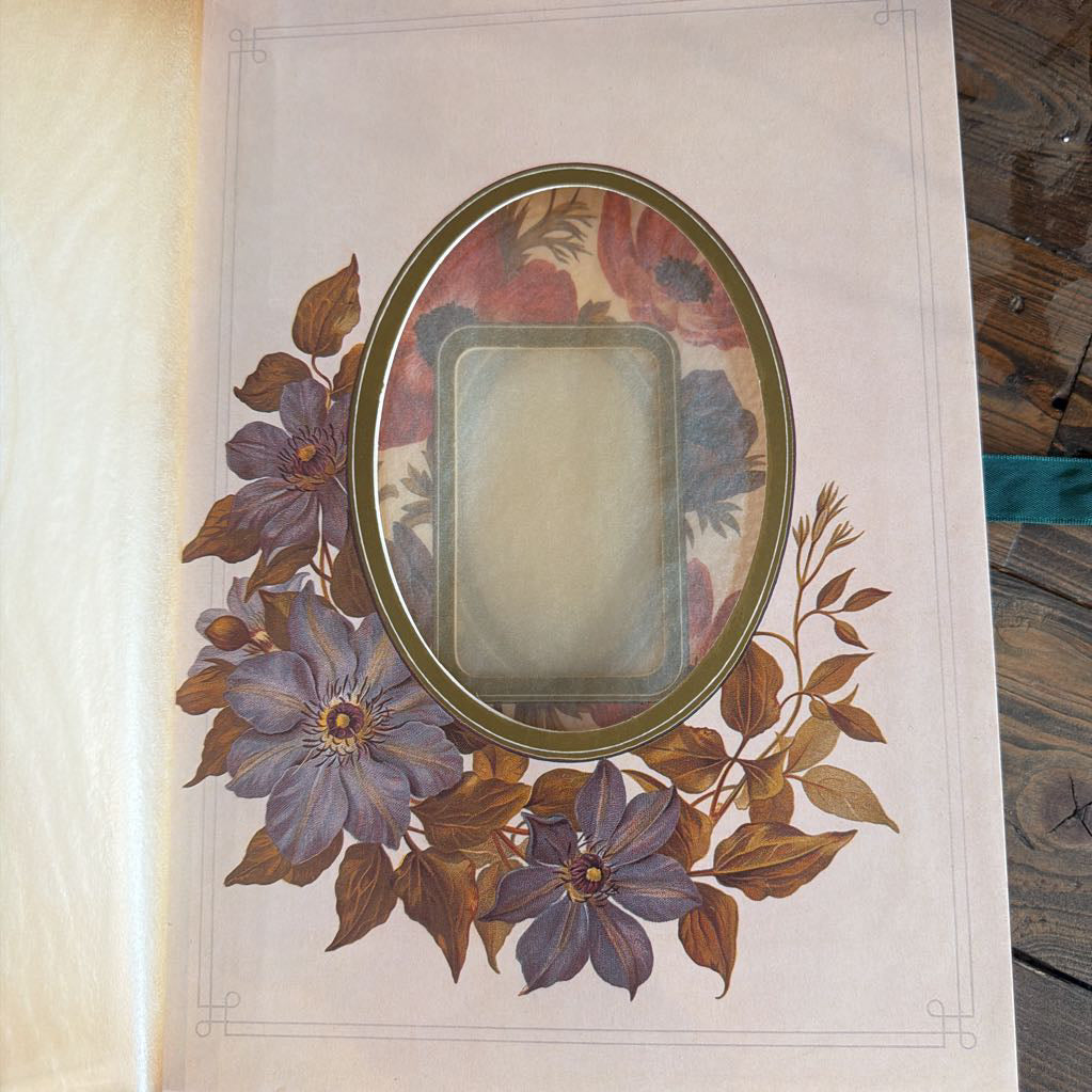An oval picture frame with a floral border design, displayed on a light beige sheet of paper. The frame features a vintage floral pattern with purple flowers and brown leaves, and the center is blanked out with a translucent protective cover.