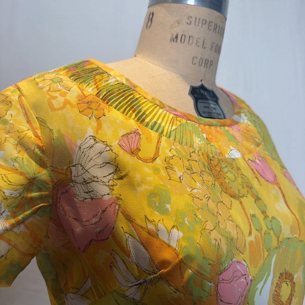 A close-up of a vintage-style blouse with a yellow floral print, displayed on a cardboard dress form. The blouse features pink, green, and white botanical motifs on a bright yellow background.