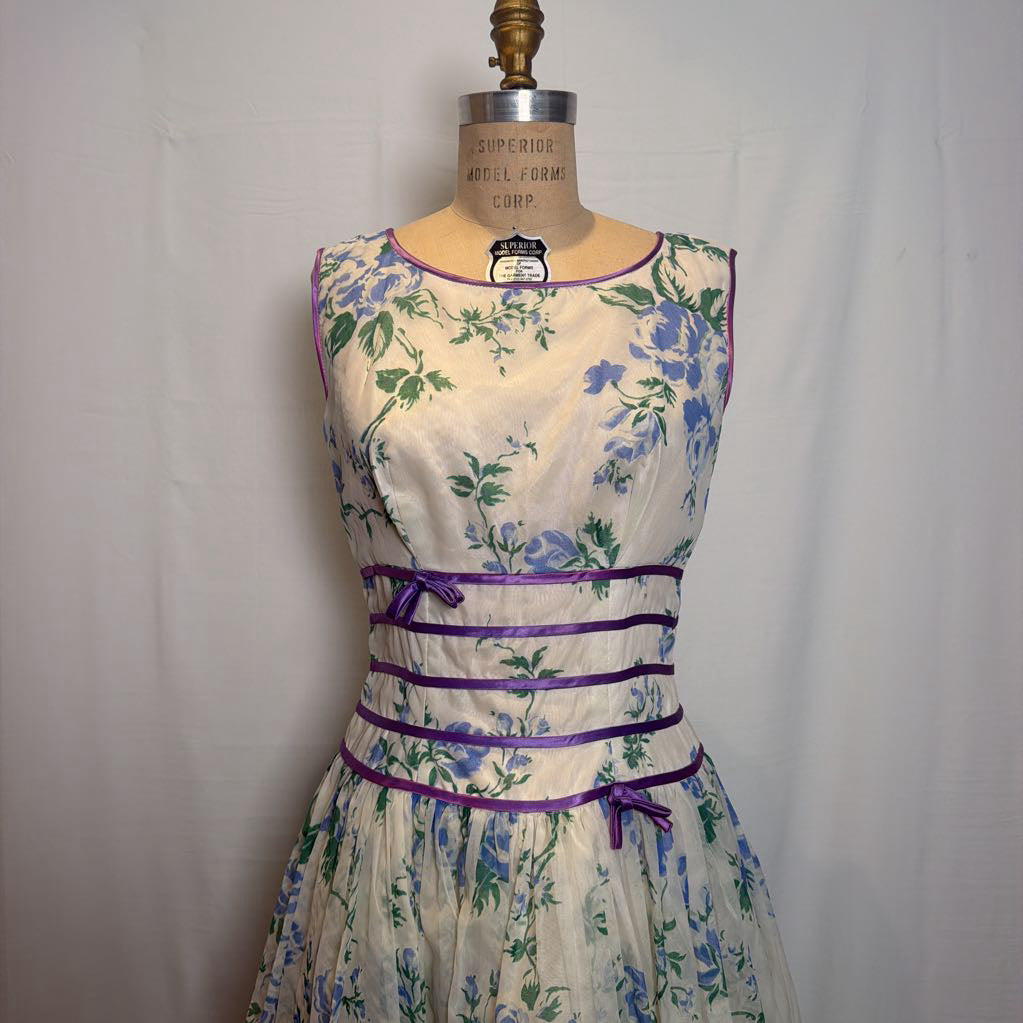 A sleeveless vintage-style A-line dress with a blue and green floral print on a cream base, featuring purple trim and waist ties, displayed on a wooden dress form.