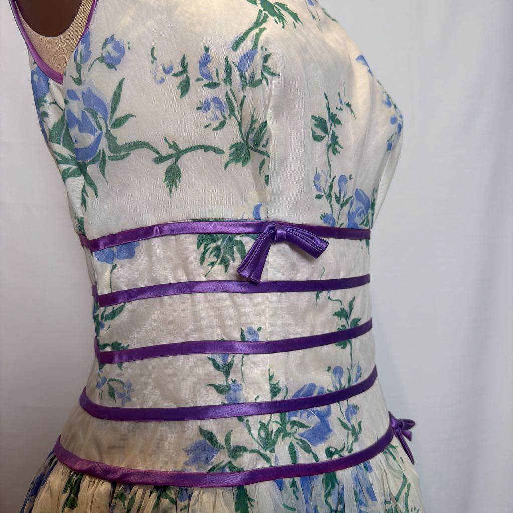 A sleeveless cream-colored dress with a blue and green floral print, featuring multiple horizontal purple satin straps and a matching purple bow at the waist.
