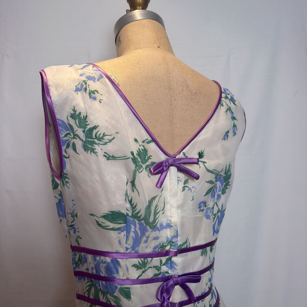 A sleeveless white dress with a blue and green floral print and purple satin trim, displayed on a vintage-style dress form from the back.