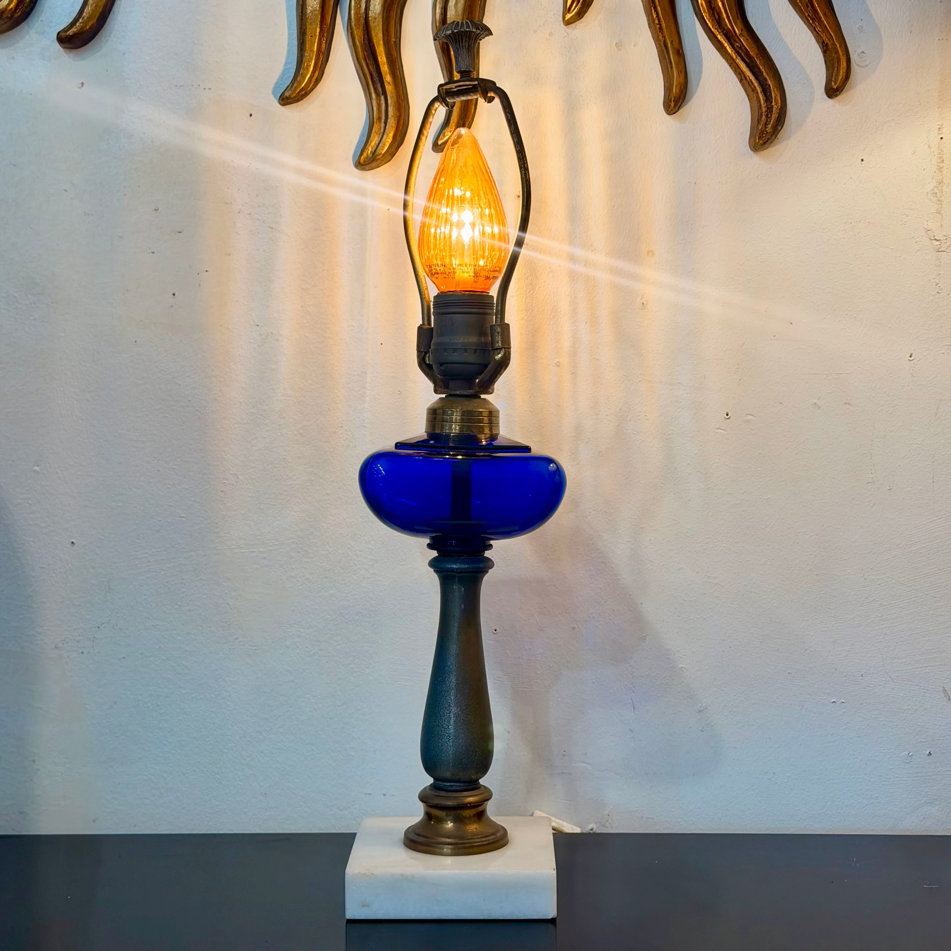 A vintage-style table lamp with a cobalt blue glass reservoir, a dark metal turned base, and a glowing warm filament bulb, set against a white wall with decorative gold-toned wall art.