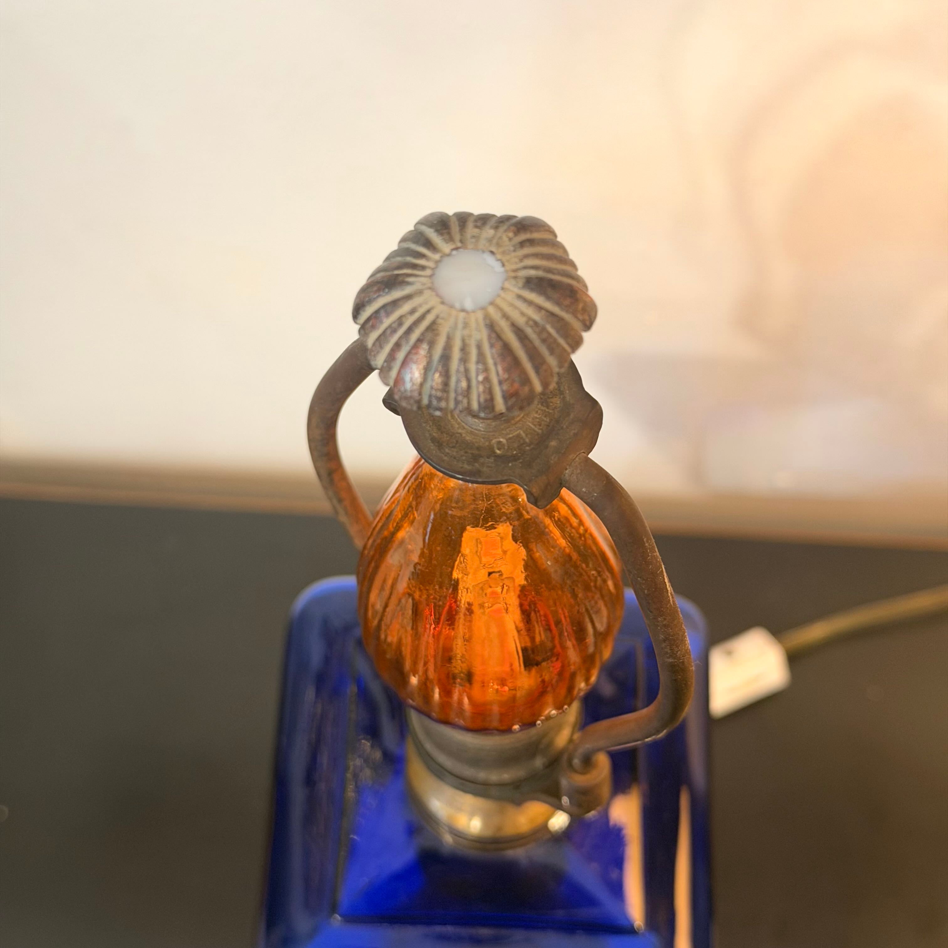 A vintage-style decorative lantern lamp with a ribbed amber glass body, dark metal side handles, and a scalloped metal top, mounted on a cobalt blue glass base.