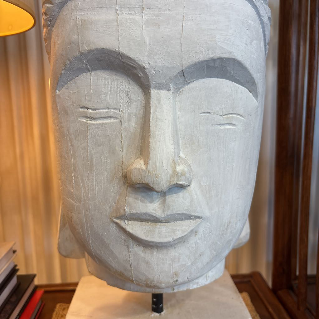 A large carved stone or concrete Buddha head sculpture with closed eyes, displayed indoors on a pedestal with a lamp and books nearby.