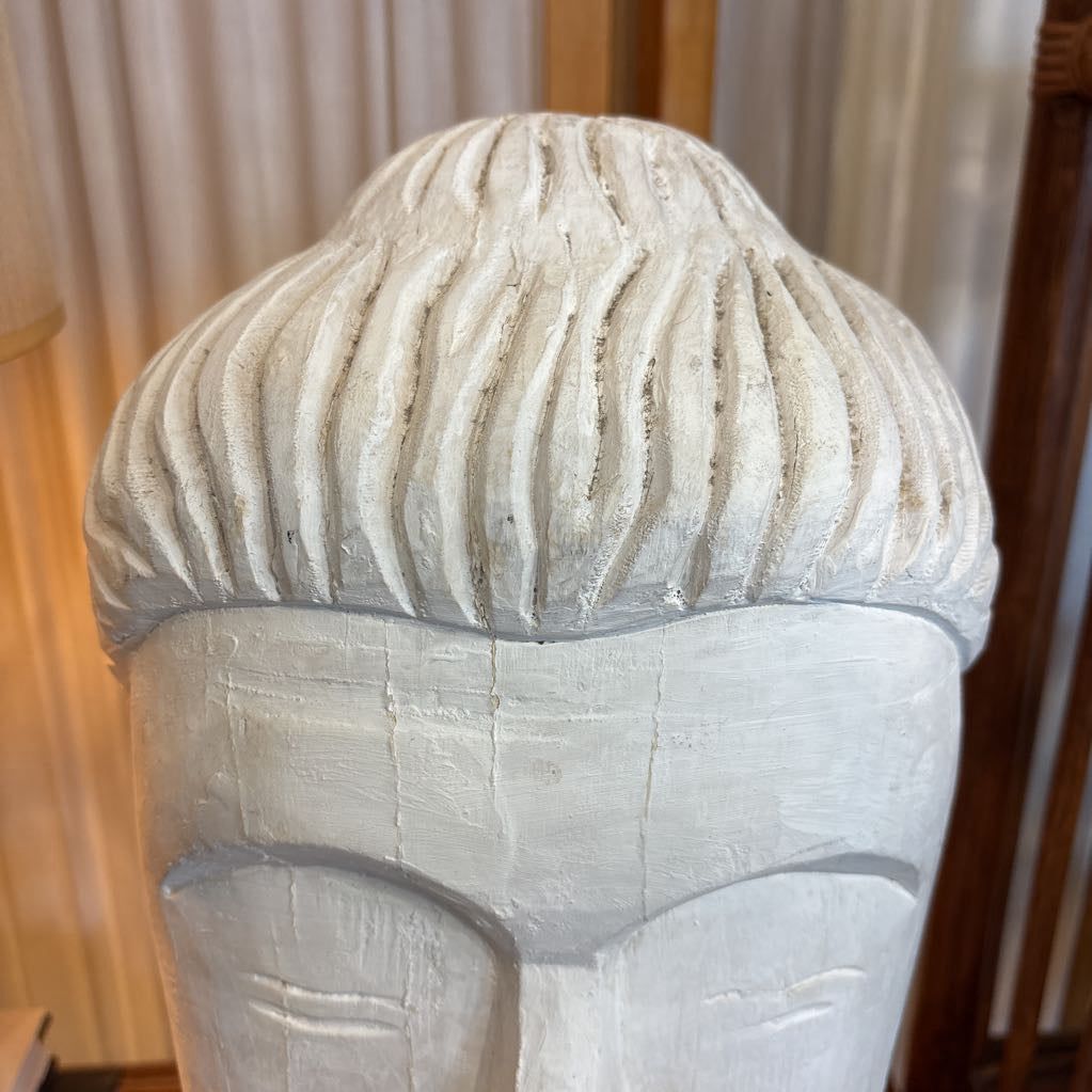 A close-up of a white textured sculpture or decorative head with carved vertical hair-like ridges, photographed indoors against a softly blurred curtain and wooden background.