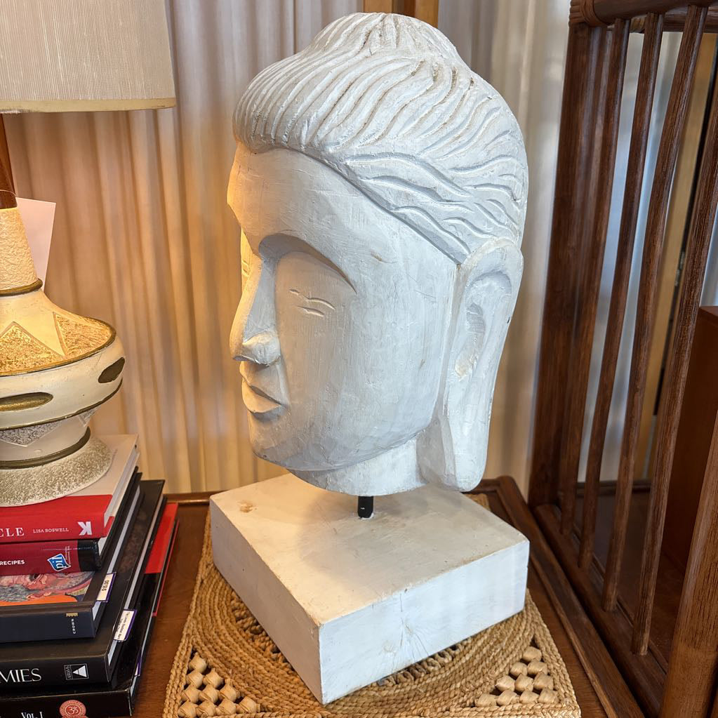 A large white carved Buddha head sculpture mounted on a matching square base, displayed on a woven mat on a wooden table in a home interior setting.