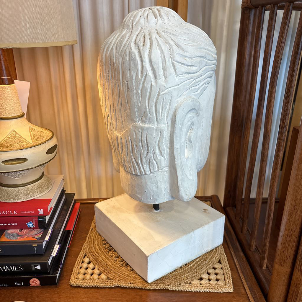 A carved white stone Buddha head sculpture mounted on a matching square base, displayed on a wooden table next to a lamp and books in an indoor setting.