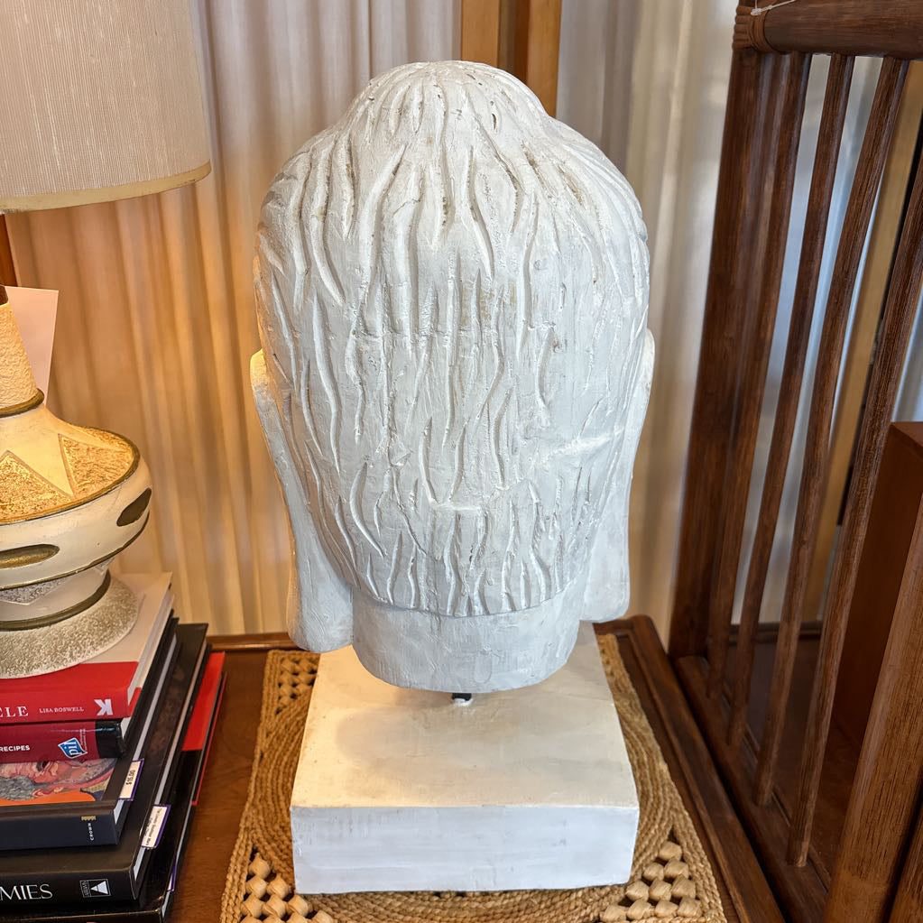 A carved white stone Buddha head sculpture mounted on a matching square pedestal, displayed in a home setting on a woven mat next to a lamp and books.