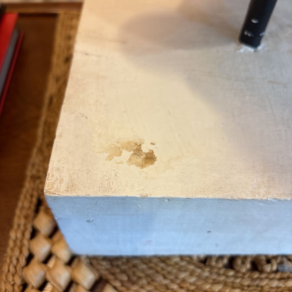 A close-up of a light beige painted wooden surface with a small brown stain or wear mark, resting on a woven wicker mat.