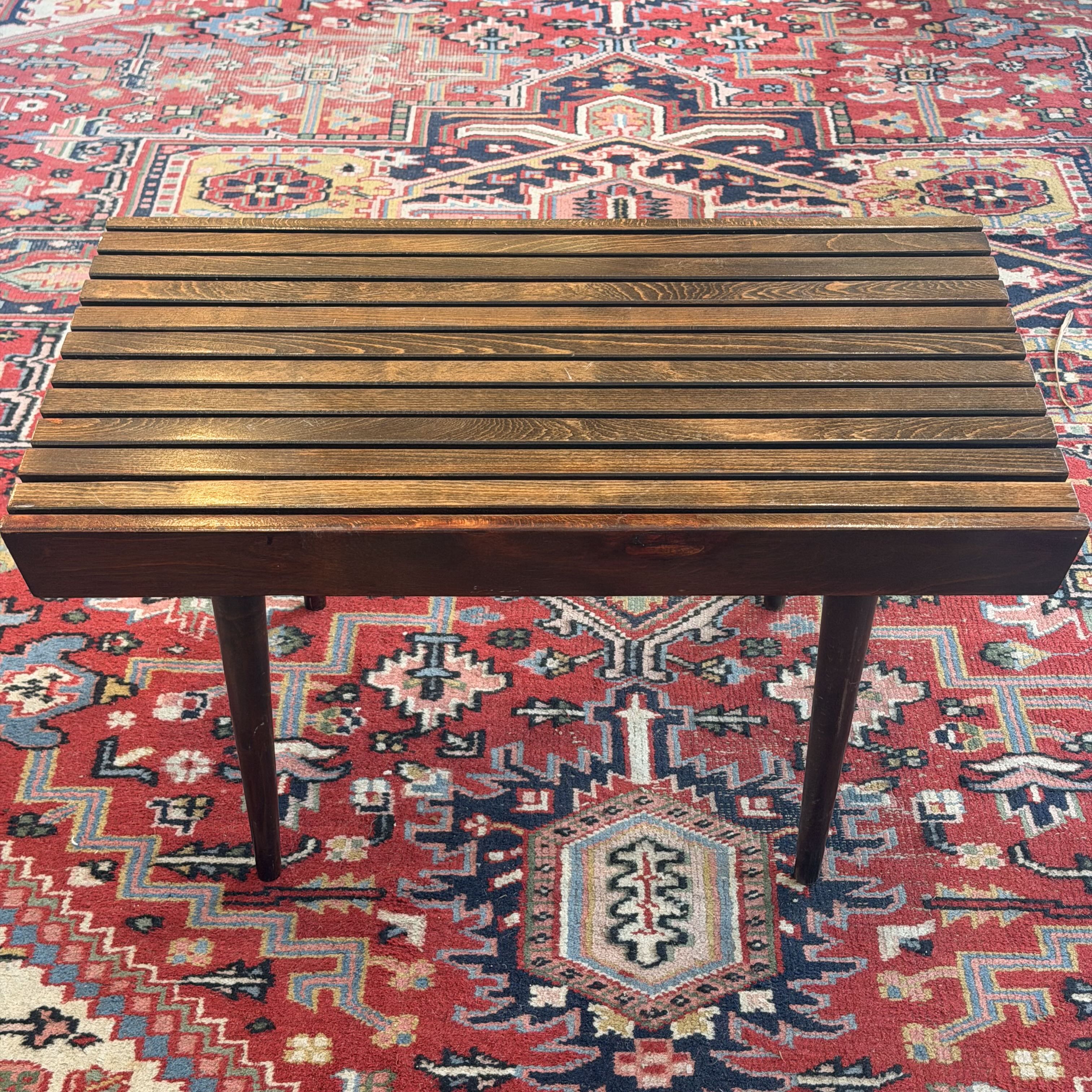 A rectangular wooden slat-top table with four legs, photographed from above on a patterned red and blue rug.