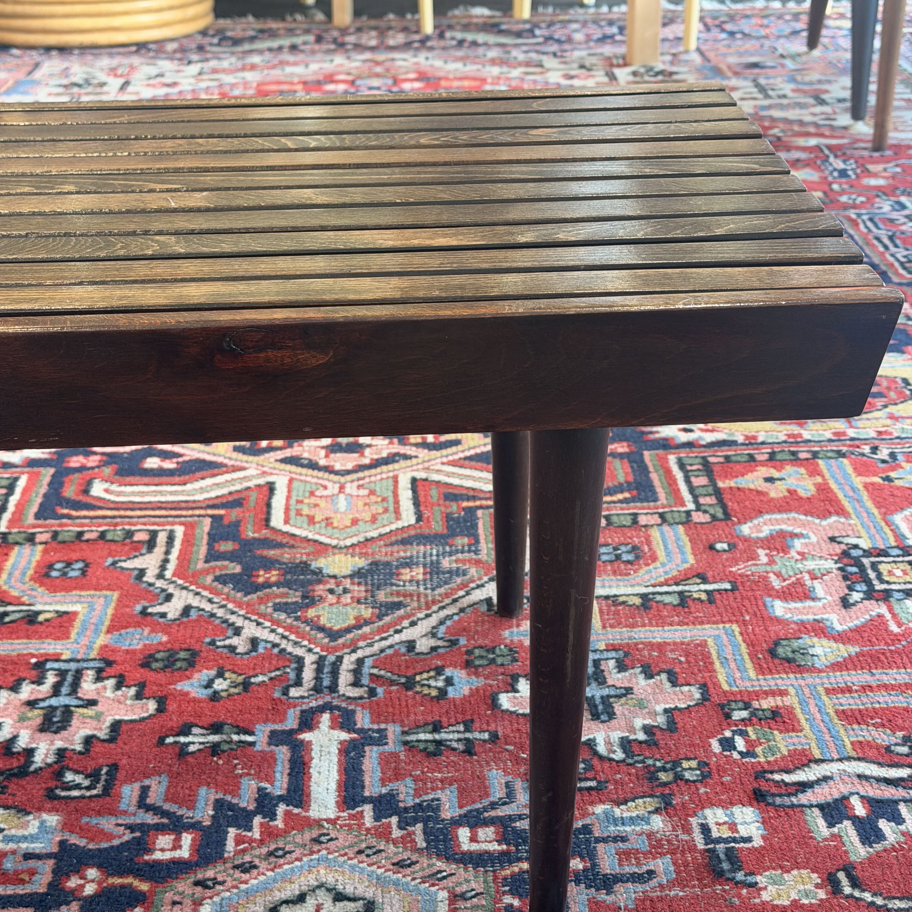 A close-up of a dark brown wooden slatted table top with matching dark legs, positioned over a red and navy patterned oriental rug.