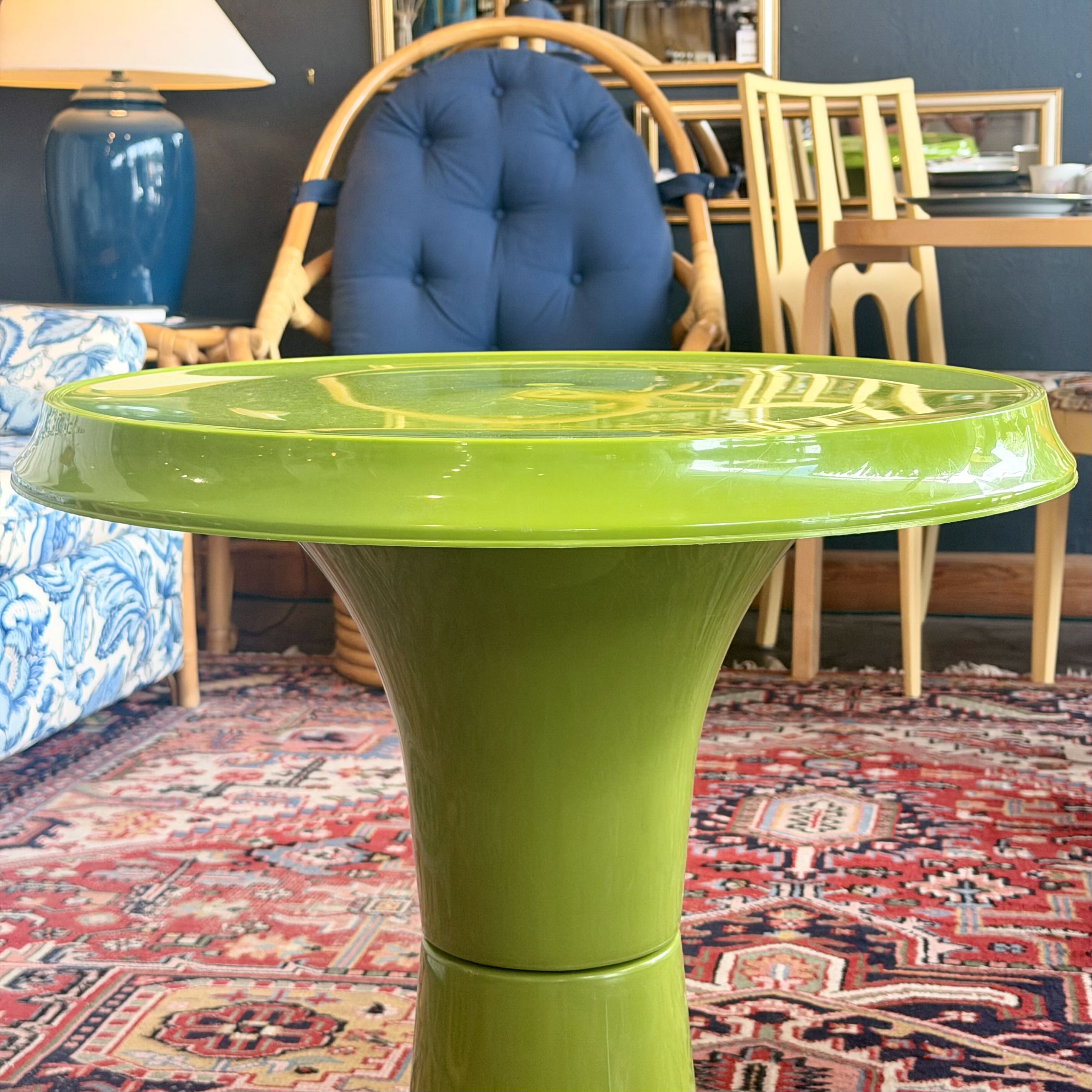 A bright lime green pedestal side table with a round top, shown in a furnished room with a patterned rug, wooden chairs, and a blue tufted chair in the background.