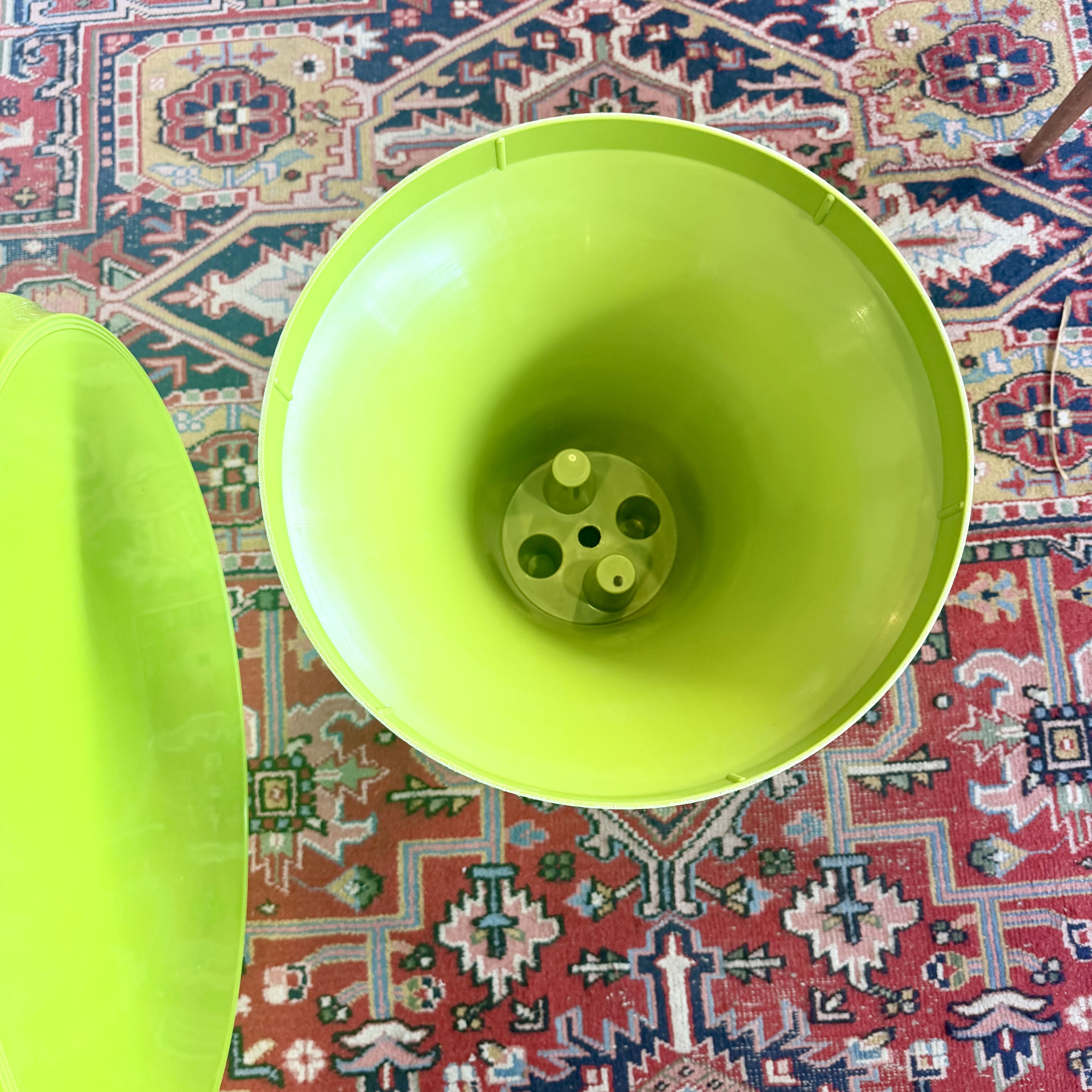 A bright lime green plastic bucket shown from a top-down view, with its matching lid placed to the left on a patterned red and blue rug.