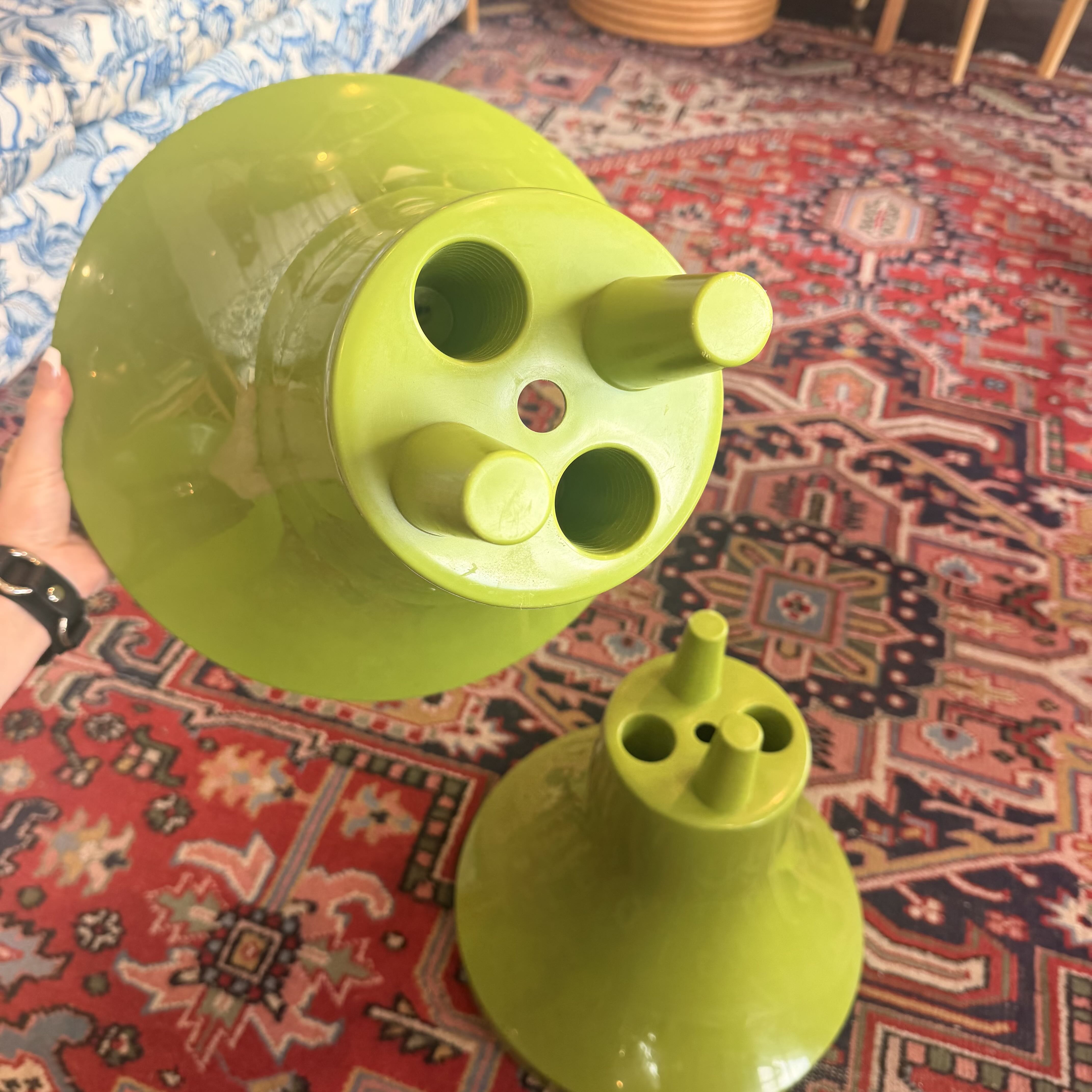 A bright lime green glass vessel, likely a teapot or coffee maker, shown disassembled with its lid and base parts held by a hand over a patterned red and blue rug.