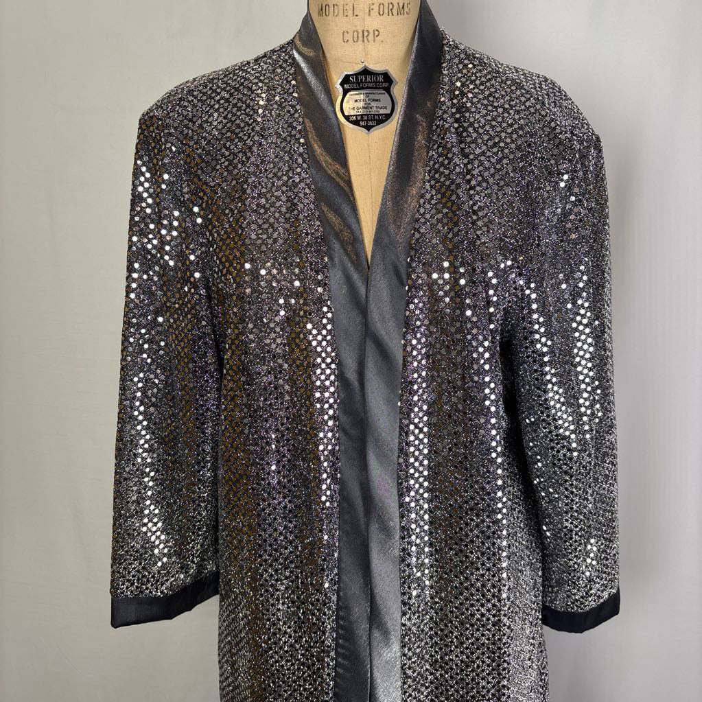 A silver sequined kimono-style jacket with black satin trim and three-quarter sleeves, displayed on a wooden dress form against a light gray background.