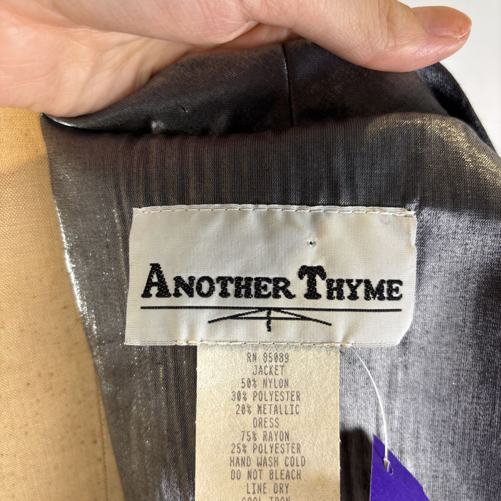 Close-up of the inside neck label of a garment held by a hand, showing the brand name 'Another Thyme' on a cream fabric tag. The garment lining is dark gray with a subtle metallic sheen, and the care instructions are printed below the brand name.