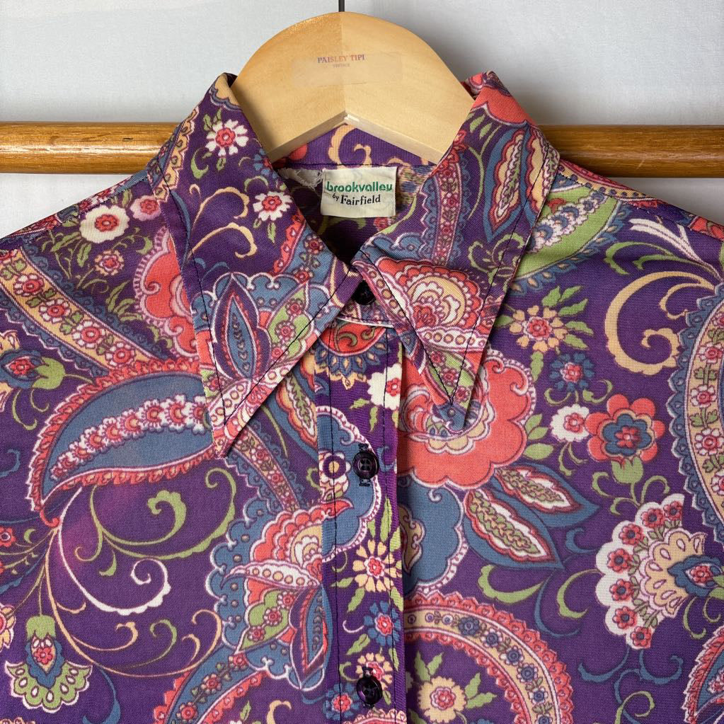 A vintage purple and paisley print button up long sleeve shirt on a hanger on a wooden rack.