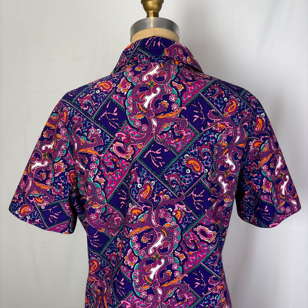 A short-sleeve button-up shirt with a vibrant purple, pink, orange, and teal paisley pattern, displayed on a dress form mannequin against a plain white background.