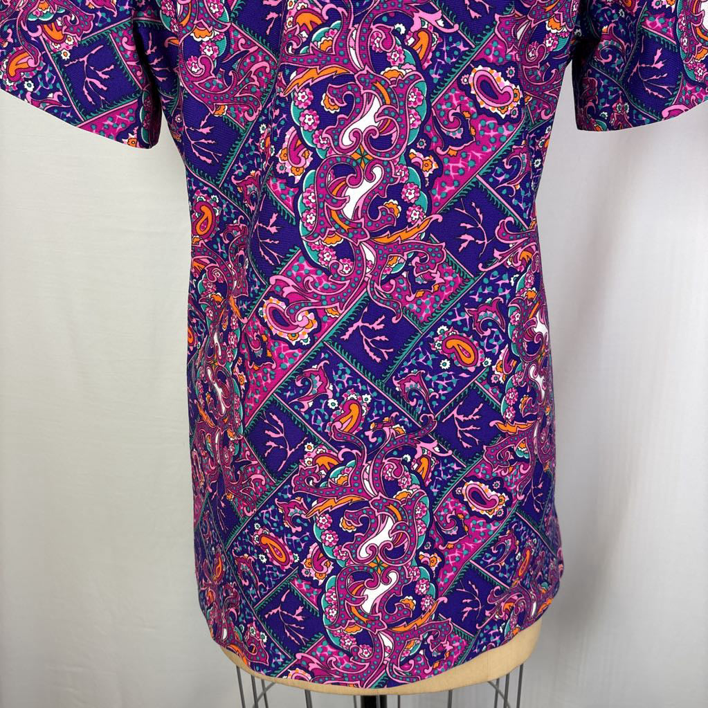 A short-sleeve button-up shirt with a vibrant purple, pink, and orange batik paisley pattern, displayed on a dress form against a white background.