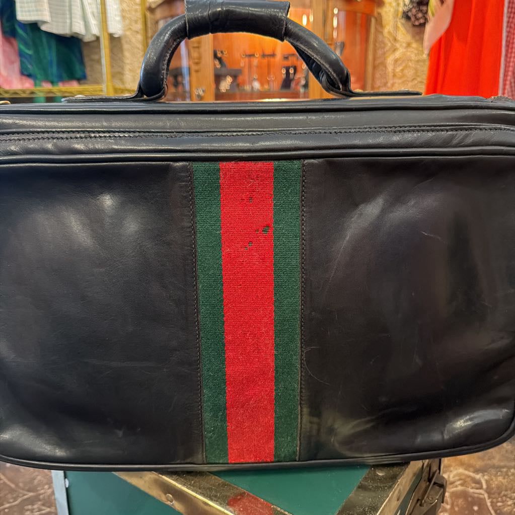 A black leather duffel bag with a green and red web stripe on the front, featuring a top handle and zippered closure, photographed in a retail store setting.