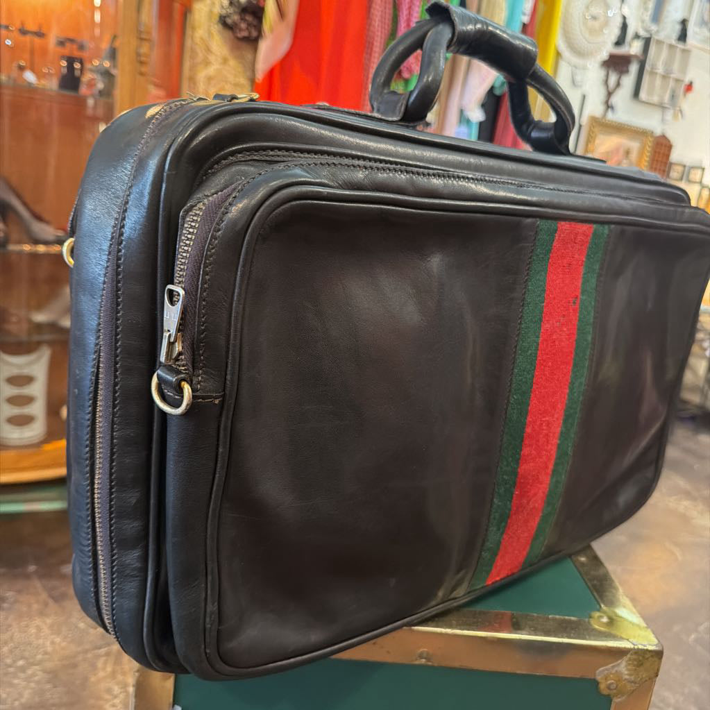 A black leather briefcase or laptop bag with a green and red web stripe detail on the front, resting on a green surface in a retail store setting.