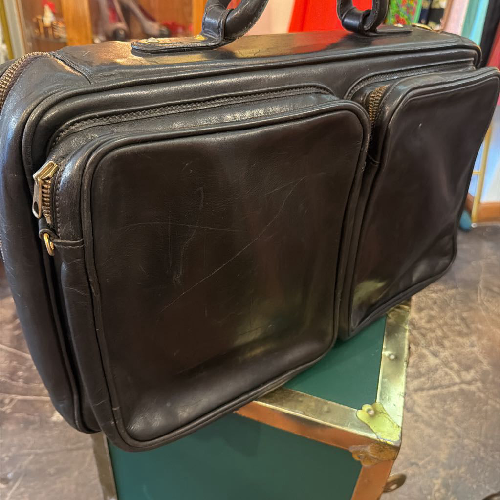 A vintage black leather briefcase or satchel with gold-tone zippers, resting on a green metal case in a shop setting.