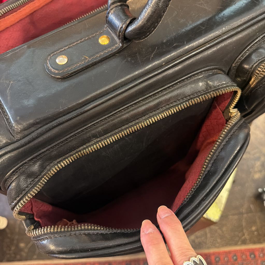Close-up of an open black leather briefcase or satchel showing a burgundy-red interior lining, with a person's fingers pointing into the compartment.