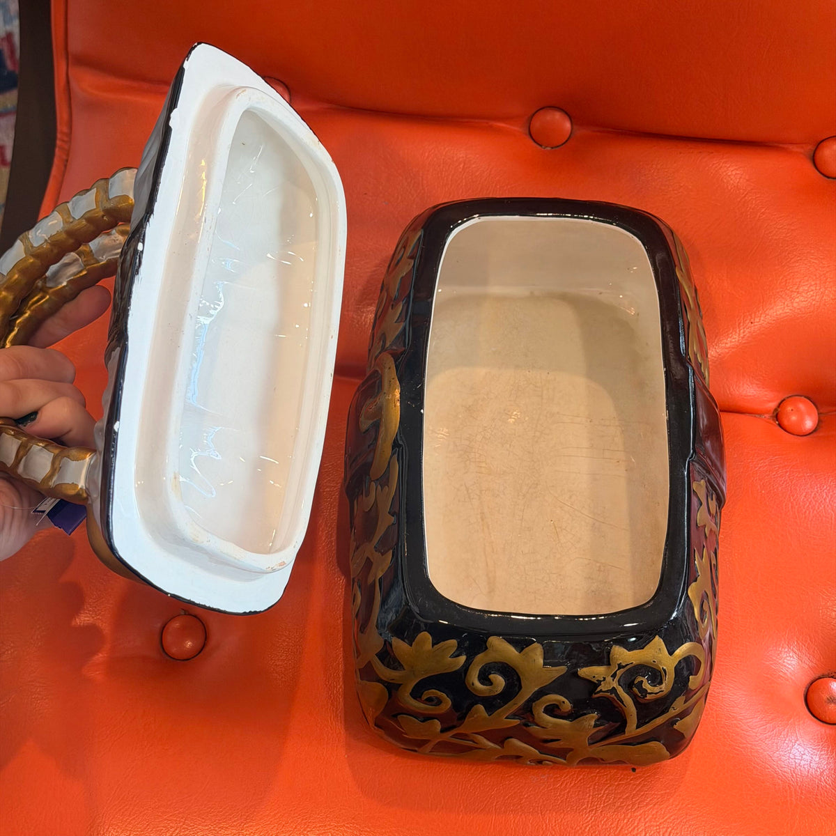 A decorative ceramic rectangular box with a hinged lid, shown open with the lid held by a hand. The box has a glossy black exterior with gold scrollwork patterns and a beige interior, set against a bright orange tufted leather background.