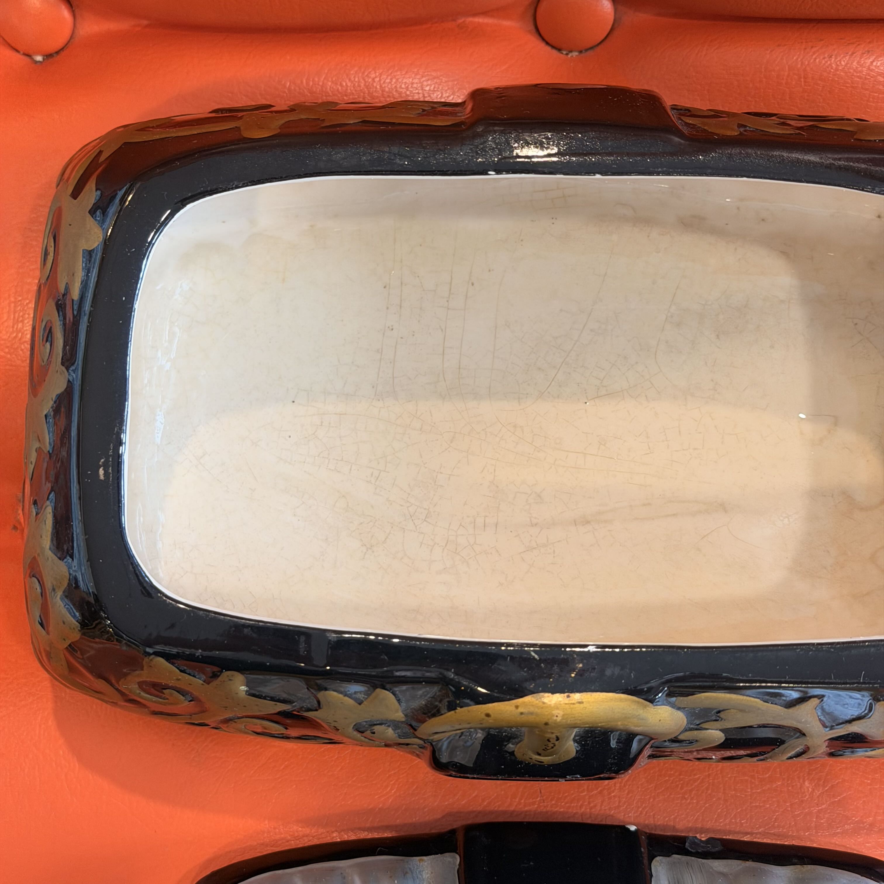 Close-up of a decorative rectangular ceramic dish or planter with a glossy black rim, cream-colored interior, and gold floral scrollwork accents, set against a bright orange background.