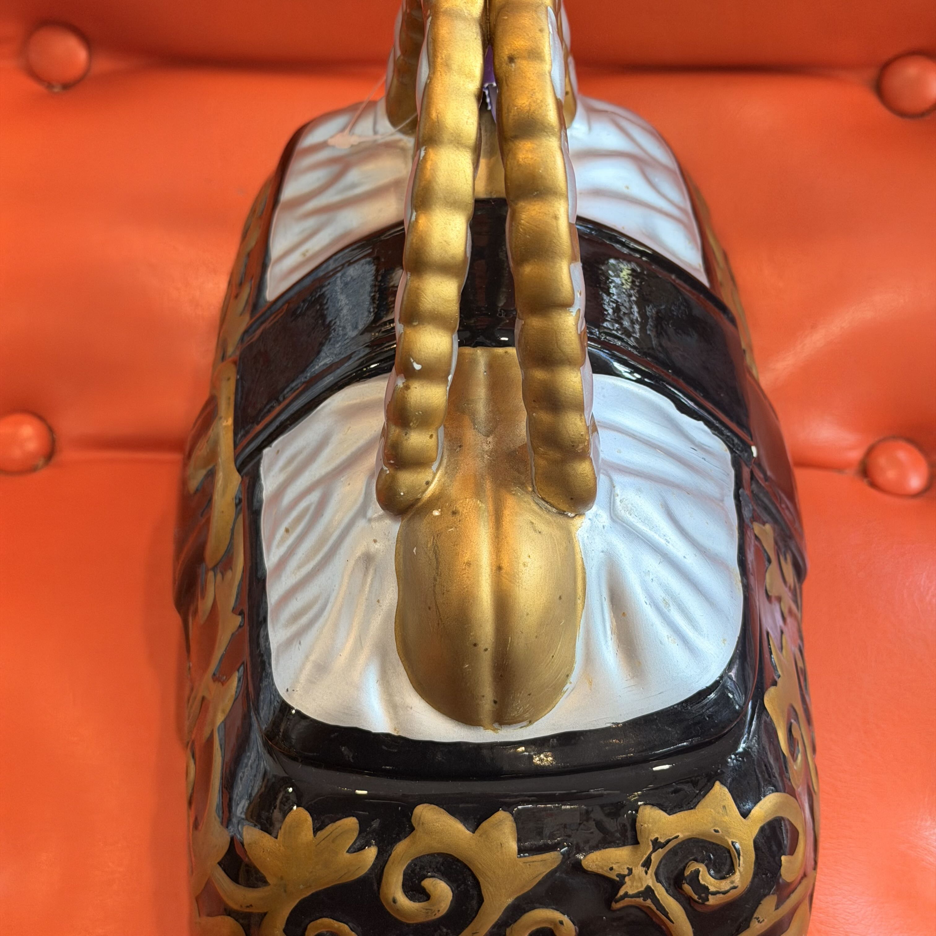 A close-up of an ornate handbag or decorative trunk with a glossy black body, white central panel, gold handles, and gold scrollwork, resting on a bright orange tufted surface.