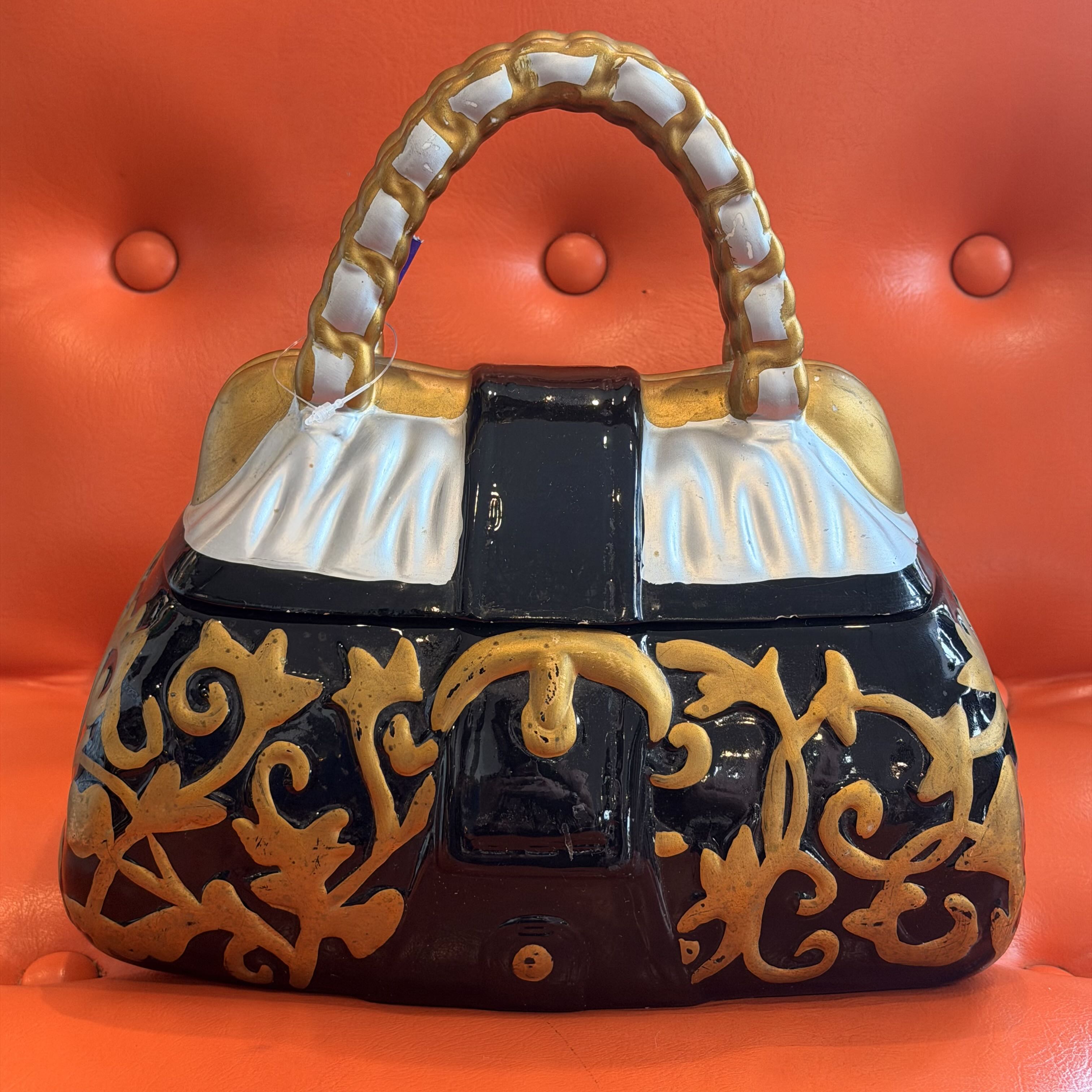 A decorative ceramic handbag-shaped object with a glossy black body, ornate gold scrollwork, white fabric-textured flap details, and a gold-and-white handle, set against a bright orange tufted background.