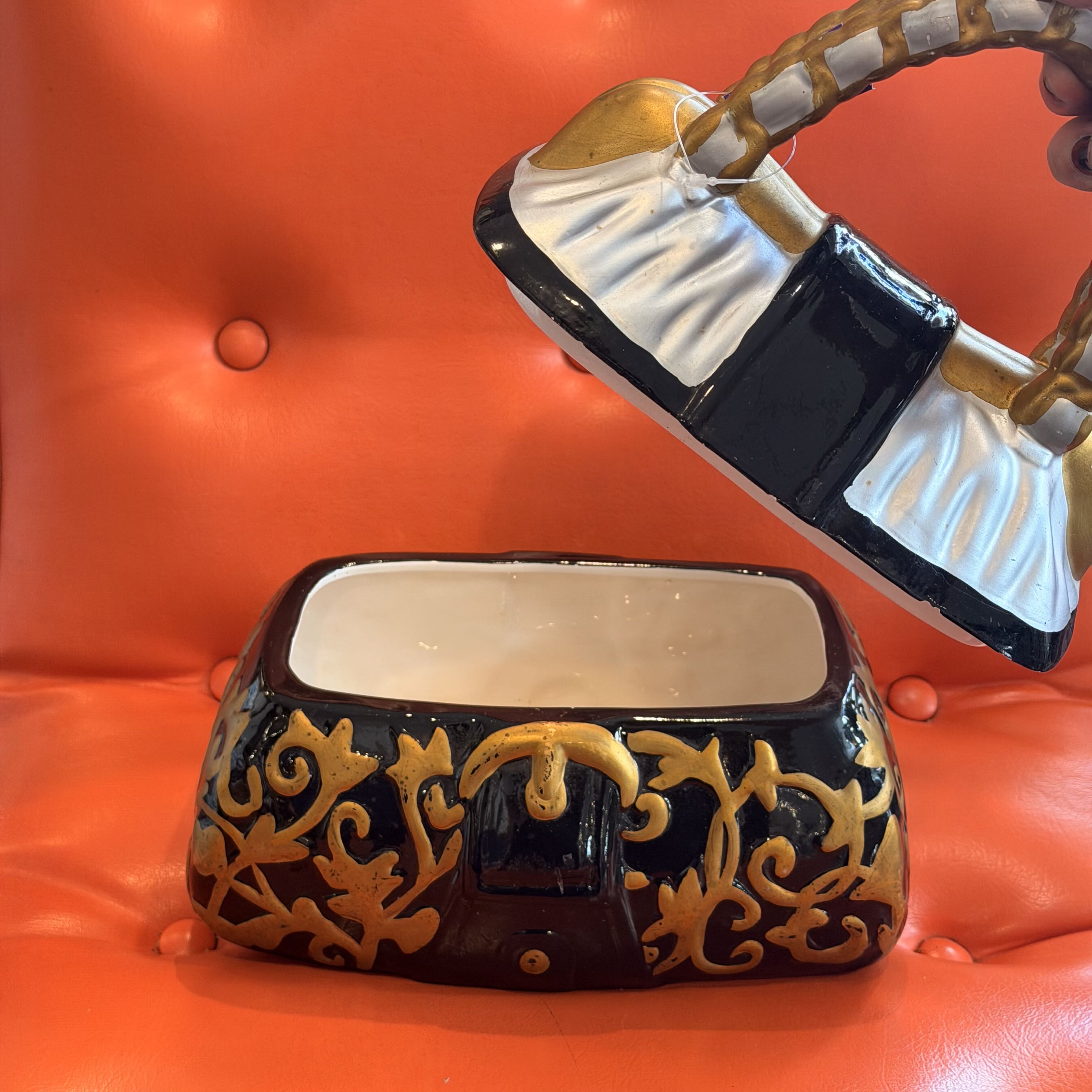 A ceramic purse-shaped container with a hinged lid, shown open against a bright orange tufted leather background. The purse is glossy black with gold scrollwork and trim, and a white fabric-like panel on the upper part of the lid.