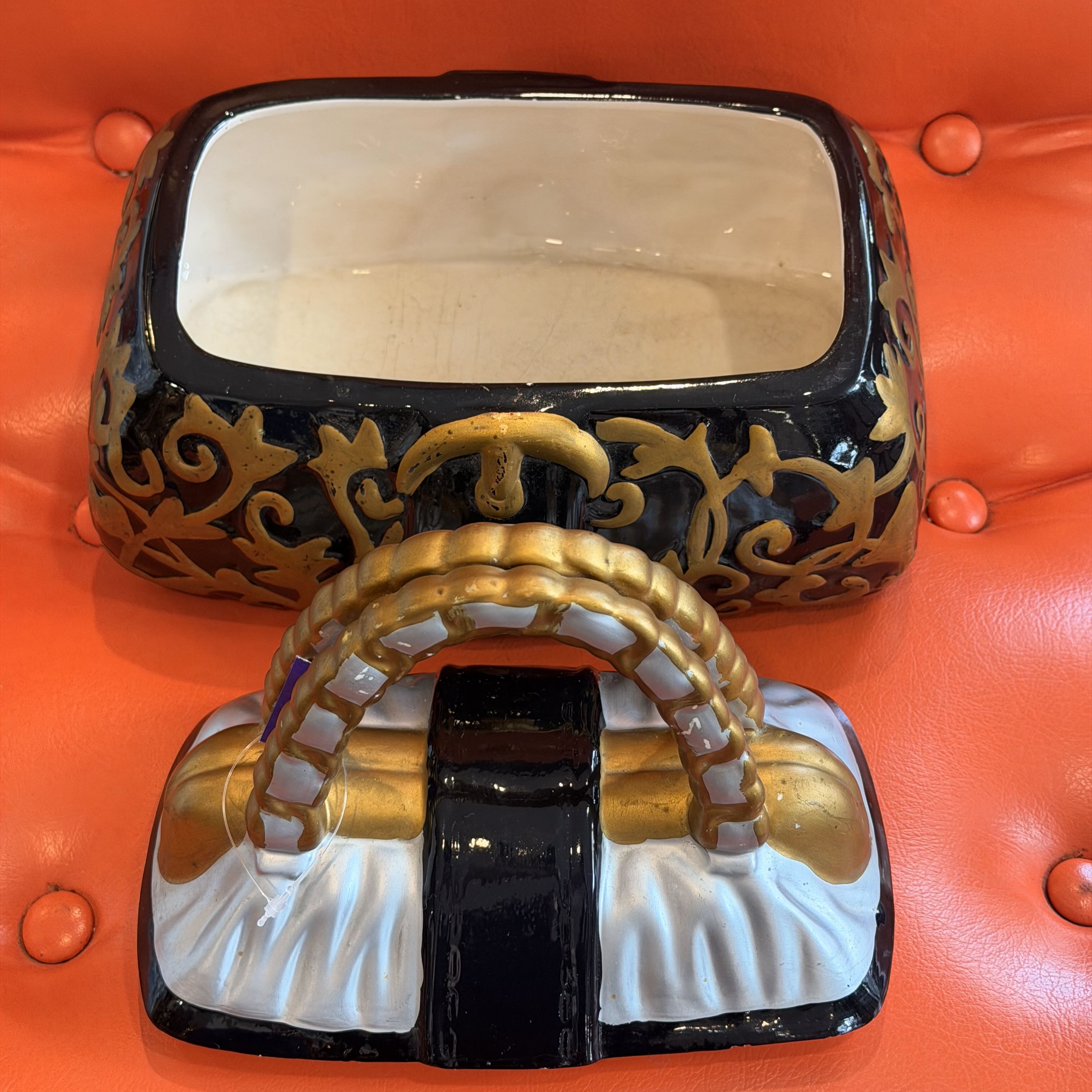 A decorative black and gold ornate box with a white interior, shown alongside a matching purse-shaped object on a bright orange tufted surface.