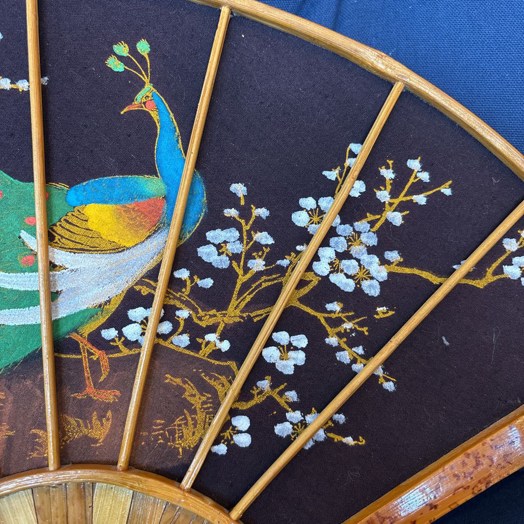 A black fabric and wooden fan wall decor with a painted peacock motif on a blue cushioned surface.