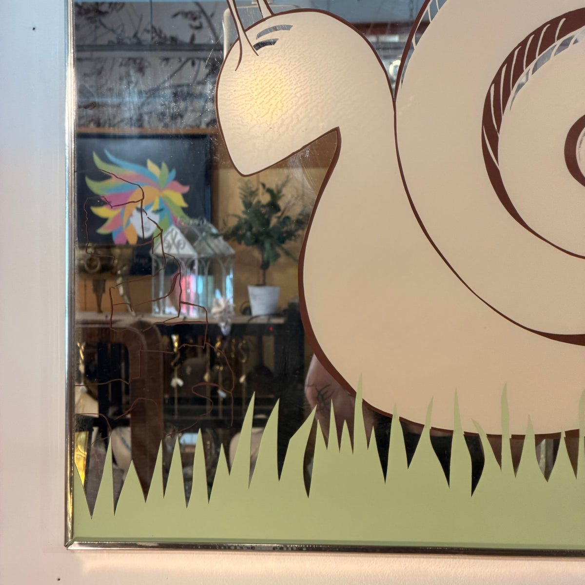 A close-up of a decorative wall mirror or glass panel featuring a stylized cream snail with brown outlines and a pale green grass silhouette at the bottom. Reflections of a room interior are visible in the glass.