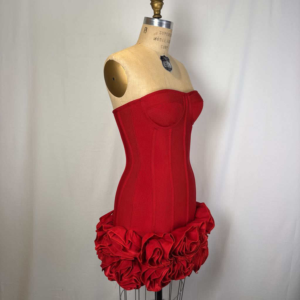 A bright red strapless corset-style dress displayed on a beige mannequin torso against a white fabric backdrop. The dress has structured boning and a ruffled rose-like skirt.