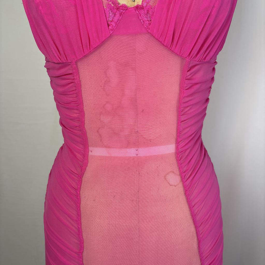 A close-up of a bright pink dress or bodysuit with ruched fabric and sheer mesh panels, shown on a mannequin or torso form against a plain light background.